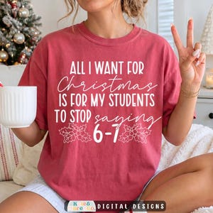 May include: A red short-sleeved t-shirt featuring white text that reads: All I Want For Christmas Is For My Students To Stop Saying 6-7, accompanied by two white holly leaf illustrations centered on the front of the garment.