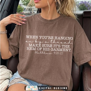 May include: A brown short-sleeved t-shirt featuring white text that reads: WHEN YOU'RE HANGING on by a thread, MAKE SURE IT'S THE HEM OF HIS GARMENT. Matthew 9:20-22. The shirt has a crew neckline and a relaxed fit with a solid brown fabric finish.