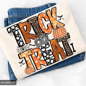 May include: A cream-colored t-shirt with the words "TRICK OR TREAT" in orange, black, and brown. The letters have various patterns, including leopard print, bats, pumpkins, and spiderwebs. The shirt is folded on top of blue jeans.