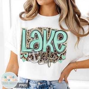 May include: A white t-shirt featuring a graphic design with the text "Lake Life" in a light teal color with a pearl-like border. The background of the text is a brown leopard print pattern, and a light gray anchor icon is positioned below the text.