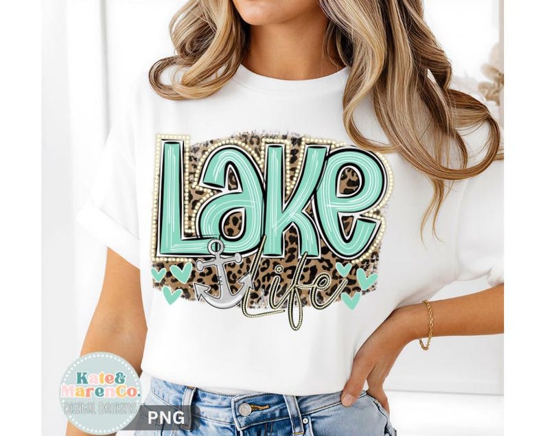 May include: A white t-shirt featuring a graphic design with the text "Lake Life" in a light teal color with a pearl-like border. The background of the text is a brown leopard print pattern, and a light gray anchor icon is positioned below the text.