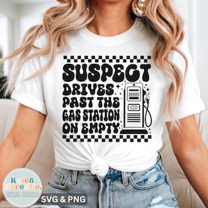 Op de afbeelding: Wit T-shirt met een zwart grafisch ontwerp. Het ontwerp toont de tekst "SUSPECT DRIVES PAST THE GAS STATION ON EMPTY" met een benzinepomp illustratie. Het shirt heeft ook een geblokte rand.