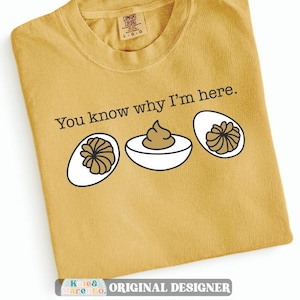 May include: A mustard yellow crew neck t-shirt featuring the black text You know why I'm here. and a graphic illustration of three deviled eggs with white egg whites and brown yolks arranged in a horizontal row across the center.