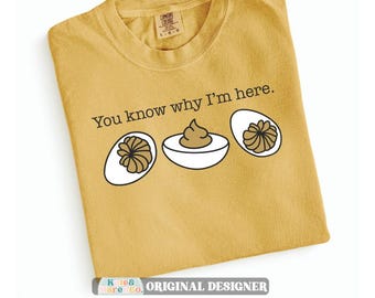 Puede incluir: Una camiseta de cuello redondo color amarillo mostaza con el texto en negro You know why I'm here. y una ilustración gráfica de tres huevos rellenos con claras blancas y yemas marrones dispuestos en una fila horizontal.