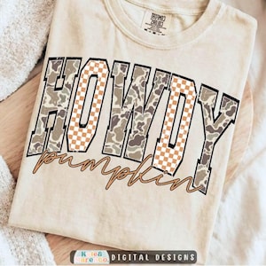 May include: A beige short-sleeve t-shirt featuring the word HOWDY in large block letters with a mix of camouflage and orange-and-white checkered patterns. Below the word HOWDY, the word pumpkin is written in a brown cursive script font.