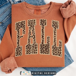 May include: A rust-colored crewneck sweatshirt featuring the word FALL in large, block-style letters with a brown and black leopard print pattern. The garment has long sleeves with ribbed cuffs and a crew neckline, layered over blue denim jeans.