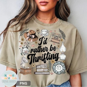 May include: Tan t-shirt with the phrase "I'd rather be thrifting" in black script. The shirt features vintage-style illustrations of a hat, teapot, telephone, books, and other antique items. The shirt is a neutral color.