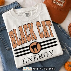 May include: A white short-sleeve t-shirt featuring the text "BLACK CAT" in large orange block letters above a circular graphic of a black cat silhouette, with three horizontal black stripes and the word "ENERGY" printed in black below.