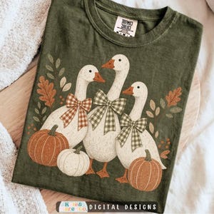 May include: An olive green t-shirt featuring a graphic of three white geese wearing plaid bows, surrounded by orange and white pumpkins and autumn leaves. The shirt has a crew neckline and a Comfort Colors brand label printed on the inside collar.
