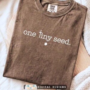 May include: A brown short-sleeved t-shirt with a crew neckline. The front features the white text "one tiny seed." with a small white dot below the word "tiny". The interior neck label reads "Comfort Colors" and indicates a size medium.