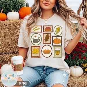 May include: A beige short-sleeve t-shirt featuring a grid of nine rectangular and circular postage stamp-style illustrations. The graphics include wheat, a bowl of apples, corn, a mug of cocoa, an oak leaf, a pumpkin, a leaf, pie, and a coffee cup.