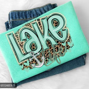 May include: A mint green t-shirt featuring a graphic print with the text "LAKE LIFE" in large, light blue letters outlined in white dots. The design includes a brown leopard print background, a gray anchor, and several small light blue hearts.