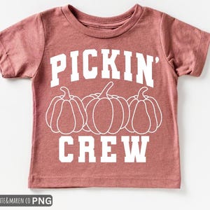 May include: A dusty rose-colored t-shirt with the words "PICKIN' CREW" in white block letters. Below the text are three white pumpkin outlines. The shirt is a short-sleeved crew neck.
