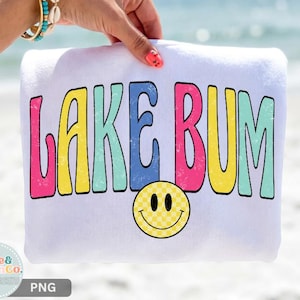 May include: A white sweatshirt featuring the text LAKE BUM in a distressed, multi-colored font with pink, yellow, blue, and teal letters. Below the text is a yellow smiley face with a checkered pattern, all printed on the front of the garment.