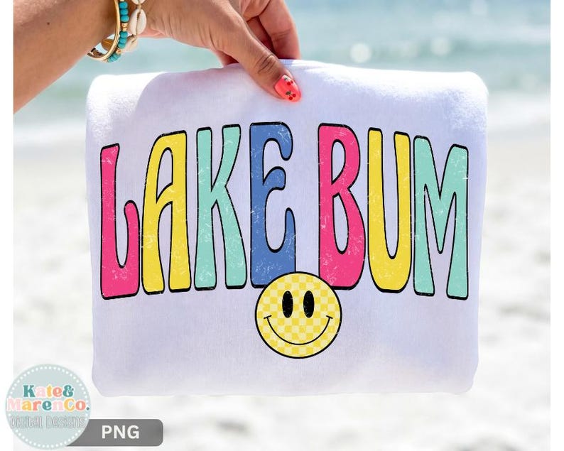 May include: A white sweatshirt featuring the text LAKE BUM in a distressed, multi-colored font with pink, yellow, blue, and teal letters. Below the text is a yellow smiley face with a checkered pattern, all printed on the front of the garment.