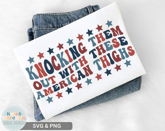 May include: White t-shirt with the text "Knocking Them Out With These American Thighs" in red, blue, and white, with star accents. The shirt is folded on top of a pair of blue jeans. The design has a retro, vintage style.