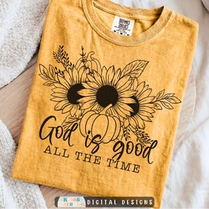 May include: A mustard yellow t-shirt featuring a black line art graphic of sunflowers and a pumpkin. The text "God is good" is written in a cursive script above the text "ALL THE TIME" in a block font. The shirt has a crew neckline.