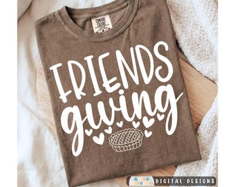 May include: A brown crew neck t-shirt featuring the white text "FRIENDS giving" in a mix of block and script fonts. Below the text is a graphic of a small pie surrounded by several small white hearts. The shirt has a Comfort Colors brand label.
