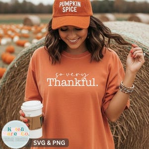 以下が含まれることがあります： 白い文字で「so very Thankful.」とプリントされたオレンジ色のTシャツと、前面に白い文字で「PUMPKIN SPICE」と刺繍されたオレンジ色の野球帽。Tシャツはクルーネックで半袖、野球帽にはカーブしたつばが付いています。
