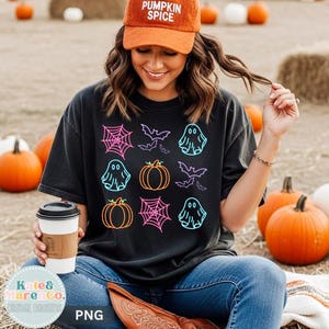 May include: A black short-sleeve t-shirt featuring a neon-colored graphic print of ghosts, pumpkins, spiderwebs, and bats. The shirt is paired with a burnt orange baseball cap with the text PUMPKIN SPICE embroidered in white on the front.