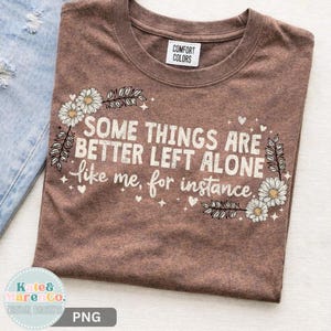 Pode incluir: Uma t-shirt castanha de gola redonda com o texto branco "SOME THINGS ARE BETTER LEFT ALONE like me, for instance." O design é emoldurado por margaridas brancas e pequenas estrelas. A t-shirt tem uma etiqueta da marca Comfort Colors.