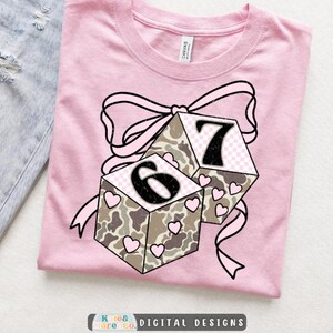 May include: A light pink t-shirt featuring a graphic of two cubes with a camouflage pattern, small pink hearts, and the numbers 6 and 7. A large pink ribbon is tied around the cubes. The design has a distressed texture and black outlines.