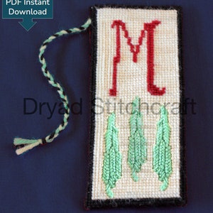 May include: A white needlepoint bookmark with a black border and a red letter "M" in the upper portion. Three green leaves are stitched in the lower portion. The bookmark has a cream and green yarn tassel.