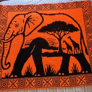 Puede incluir: Una manta de ganchillo naranja y negra con un elefante, un árbol y jirafas. La manta tiene un borde decorativo con flecos. El diseño representa una escena de la sabana africana.