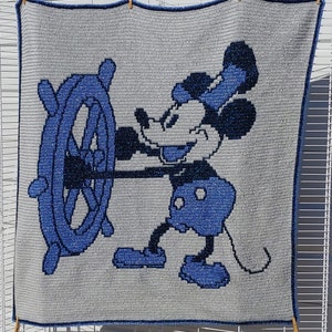 Puede incluir: Una manta de crochet azul y blanca con Mickey Mouse sosteniendo un timón. La manta tiene un fondo gris y detalles azules.