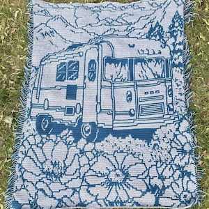 Peut inclure: Une couverture en crochet avec un motif bleu et blanc représentant un camping-car avec des montagnes et des fleurs. La couverture a une bordure frangée.
