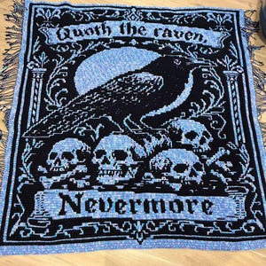 Könnte beinhalten: Eine rechteckige Häkeldecke mit einem schwarz-hellblauen Design eines Raben, der auf Totenköpfen sitzt. Der Text "Quoth the raven" steht oben und "Nevermore" unten in einer gotischen Schriftart. Die Decke hat an den Rändern Fransen.