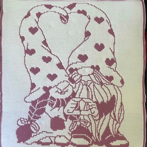 May include: A crocheted cushion featuring a pink and white design of two gnomes holding hearts. The gnomes are wearing hats with hearts on them.