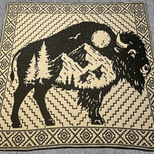 May include: A black and cream-colored crocheted blanket featuring a bison silhouette with a mountain and forest scene. The blanket has a geometric border and a zig-zag pattern. The bison design includes a moon and stars.