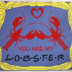 Op de afbeelding: Een gehaakt deken met een blauwe achtergrond en een gele rand. Het deken heeft twee rode kreeften die elkaar aankijken met een hart erboven. De tekst "YOU ARE MY L-O-B-S-T-E-R" staat in het zwart onder de kreeften.