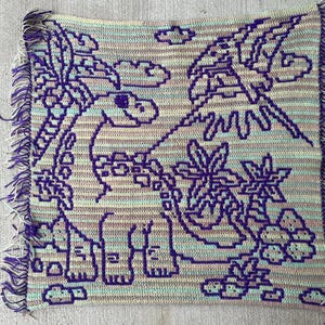 May include: A handmade crochet blanket with a dinosaur design in purple, blue, and beige. The blanket has a fringed edge and depicts a dinosaur, palm trees, and clouds. The blanket is approximately 100 cm by 100 cm.