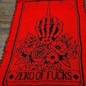 May include: A red crocheted blanket with a black design. The design features a skeleton hand giving the middle finger, roses, and the text "ZERO OF FUCKS". The blanket has a fringed edge.