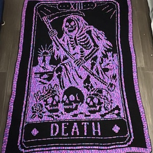 Könnte beinhalten: Eine rechteckige Häkeldecke mit einem Tod-Tarotkarten-Motiv. Der schwarze Hintergrund bildet einen Kontrast zu dem leuchtend violetten und blauen melierten Garn, das für das Skelett mit Sense, drei Totenköpfe und das Wort DEATH verwendet wurde.