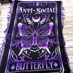 May include: A black and purple crocheted blanket featuring the words "ANTI-SOCIAL" and "BUTTERFLY" in white. The design includes a butterfly motif. The blanket has fringed edges and is made from soft yarn.