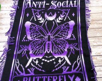 May include: A black and purple crocheted blanket featuring the words "ANTI-SOCIAL" and "BUTTERFLY" in white. The design includes a butterfly motif. The blanket has fringed edges and is made from soft yarn.