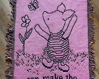 May include: Pink crocheted blanket with a black and white image of a bear holding a flower. The blanket has fringe around the edges and the text "Sometimes the smallest creature can make the biggest impact".