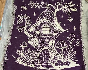 May include: A crocheted throw blanket with a purple background and a white design of a tree house with a window and a door. The tree house is surrounded by leaves, mushrooms, and stars.