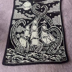 Könnte beinhalten: Schwarz-weiße Häkeldecke mit einem Schiff, das von einem riesigen Tintenfisch angegriffen wird. Das Design umfasst einen Vollmond, Wolken und Wellen. Die Decke hat einen schwarzen Rand und ist aus weichem Garn gefertigt.