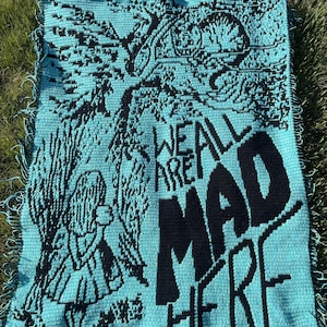 May include: A rectangular crochet blanket in turquoise and black featuring a graphic of a person standing under a tree with a cat in the branches. The text "WE ARE ALL MAD HERE" is prominently displayed in large black letters on the right side.