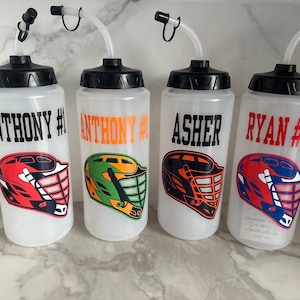 May include: Four clear plastic water bottles with black lids and straws. Each bottle has a different name and number printed on it, along with a lacrosse helmet graphic. The names are Anthony #1, Anthony #1, Asher, and Ryan #18.