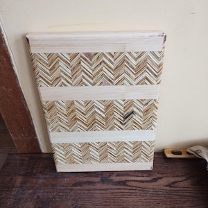 May include: A rectangular wooden wastebasket with a chevron pattern. The design features alternating rows of light and dark wood pieces, creating a textured surface. Horizontal bands of lighter wood separate the chevron sections.