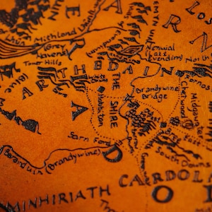 May include: A close-up of a leather map of Middle-earth from The Lord of the Rings. The map is brown with black ink and features the Shire, the Brandy Wine River, and the surrounding areas.