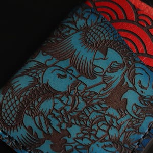 May include: A blue leather wallet with a detailed embossed design of koi fish and waves. The wallet is stitched with brown thread and has a red leather panel with a wave pattern.