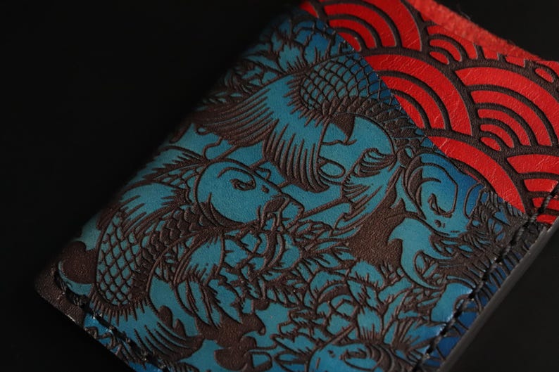 May include: A blue leather wallet with a detailed embossed design of koi fish and waves. The wallet is stitched with brown thread and has a red leather panel with a wave pattern.