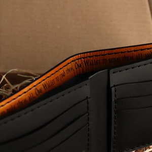 May include: A black leather wallet with an orange leather trim. The inside of the wallet has a quote that reads "One Wallet to find them, One Wallet to bring them all and in the...".