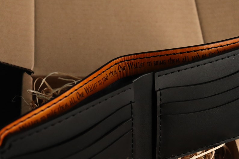 May include: A black leather wallet with an orange leather trim. The inside of the wallet has a quote that reads "One Wallet to find them, One Wallet to bring them all and in the...".